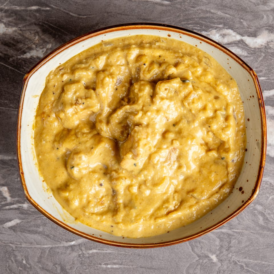 Overhead view of a bowl of creamy, yellow Indian curry with visible spices on a marble surface.